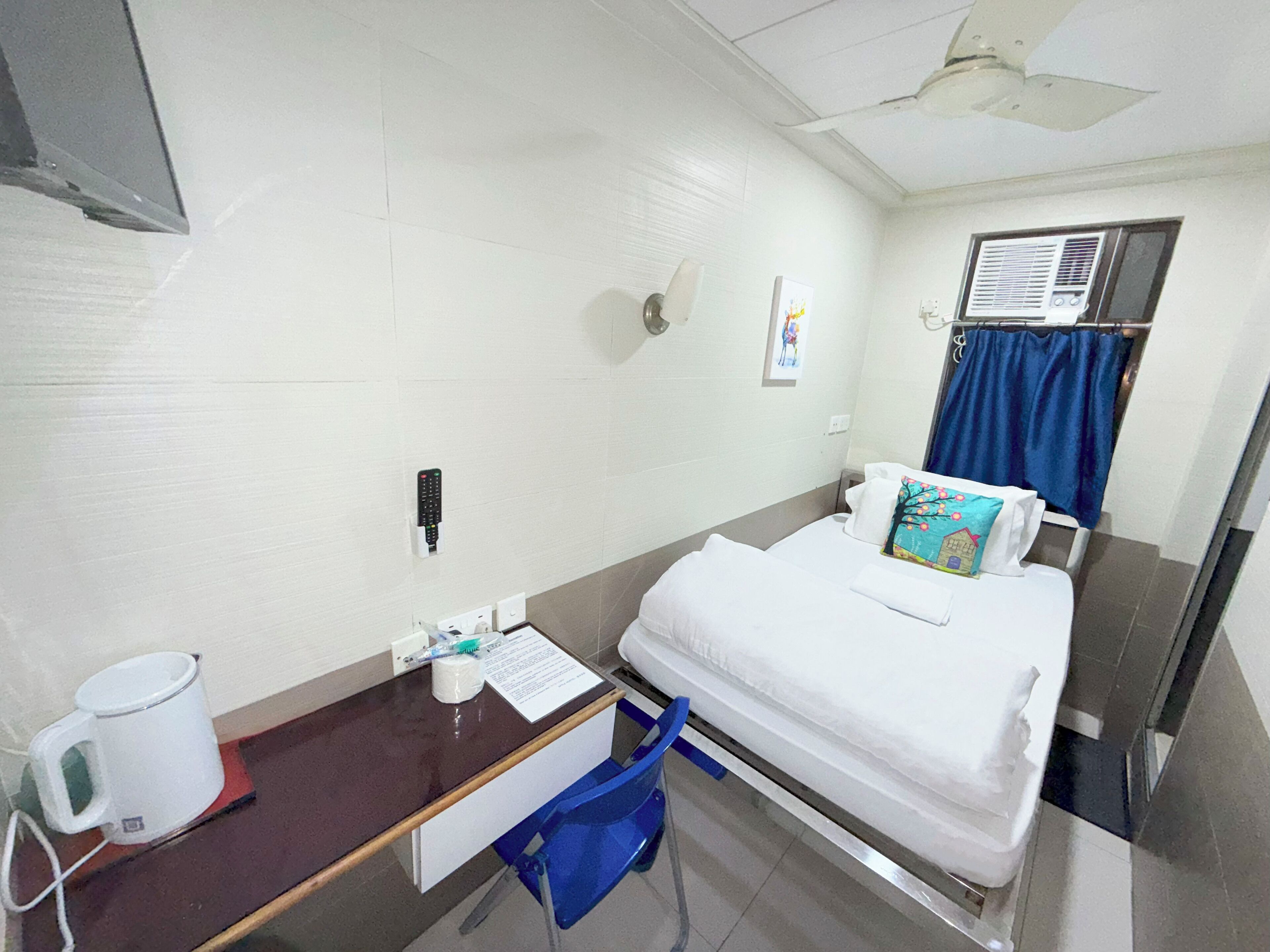 A hotel room with a bed, a desk with a cup and tissues, a blue chair, a window with a curtain, and a ceiling fan.
