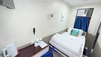 A hotel room with a bed, a desk with a cup and tissues, a blue chair, a window with a curtain, and a ceiling fan.