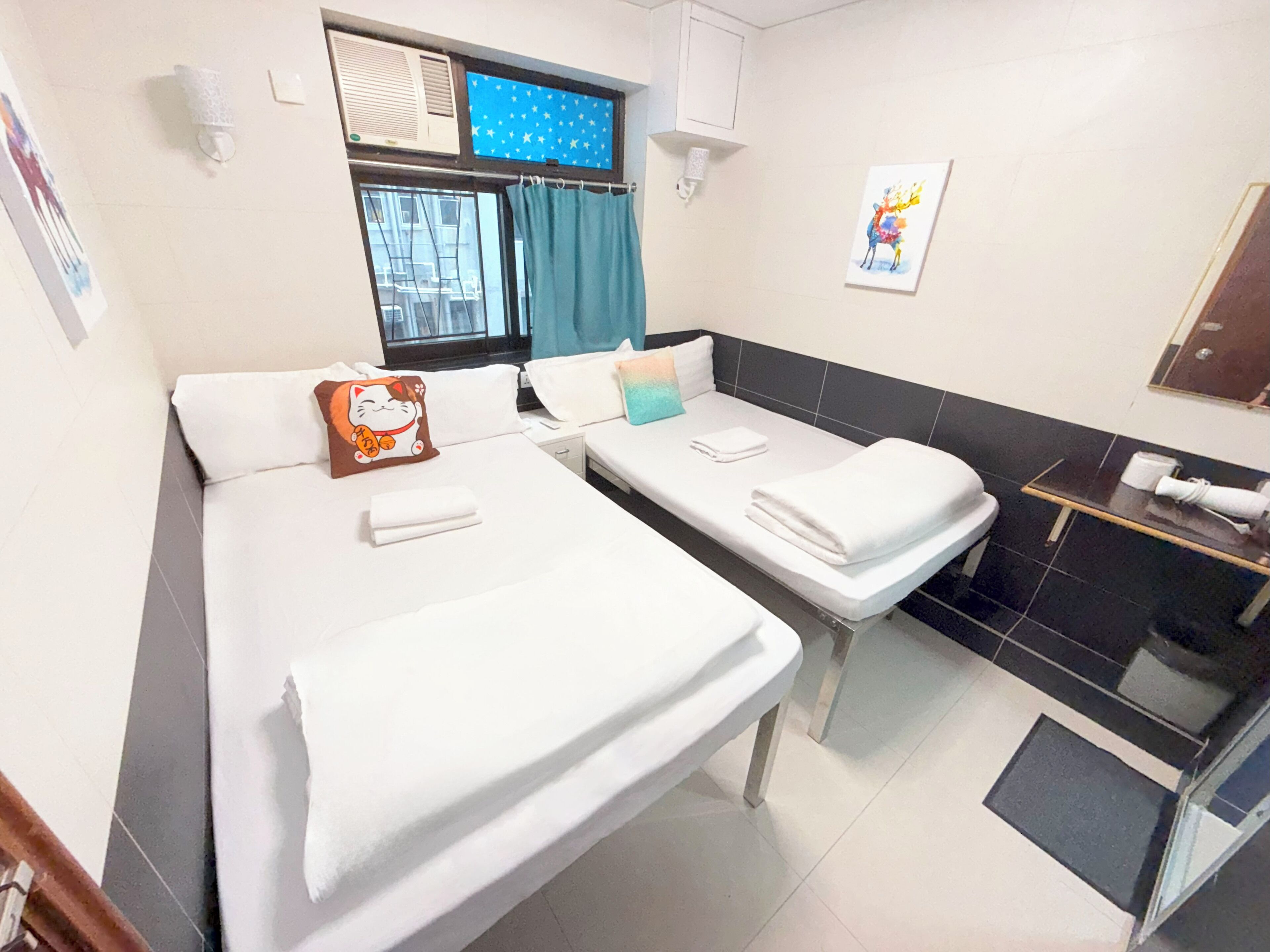 A hotel room with two beds, a small kitchenette, a window with curtains, and a painting on the wall.