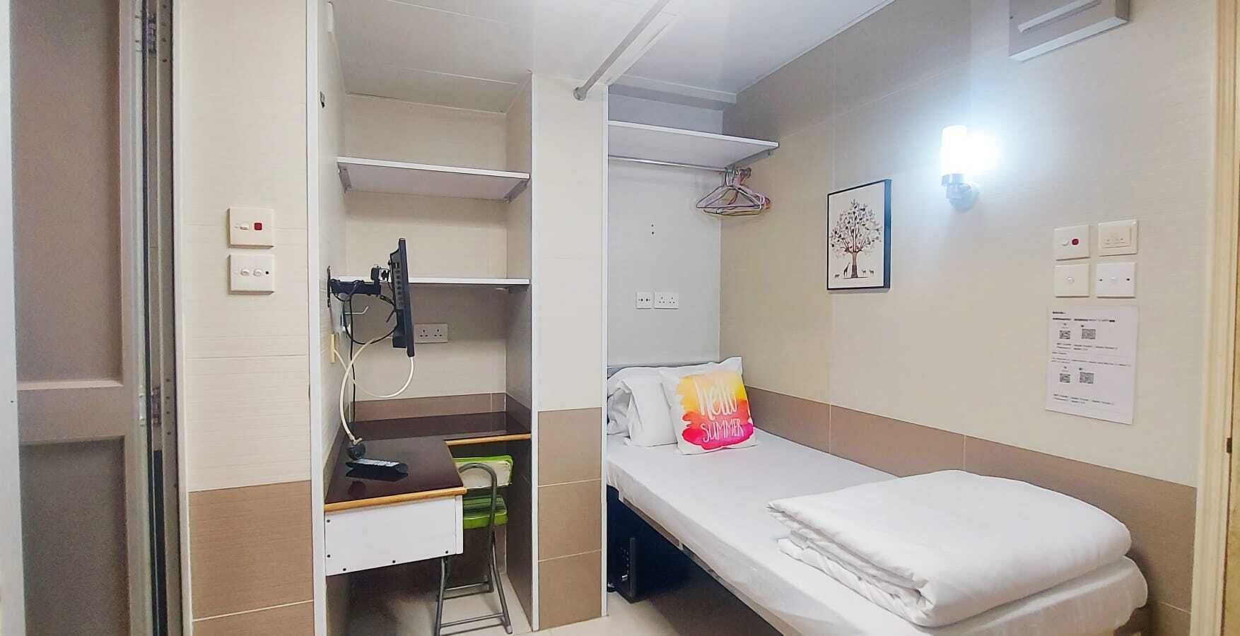 A small, compact room with a single bed, a desk with a chair, a shelf, and a wall-mounted heater.