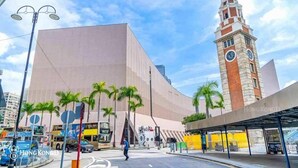 A modern building with a prominent clock tower, palm trees, and a covered bus stop. - Apple Hostel (Kowloon)