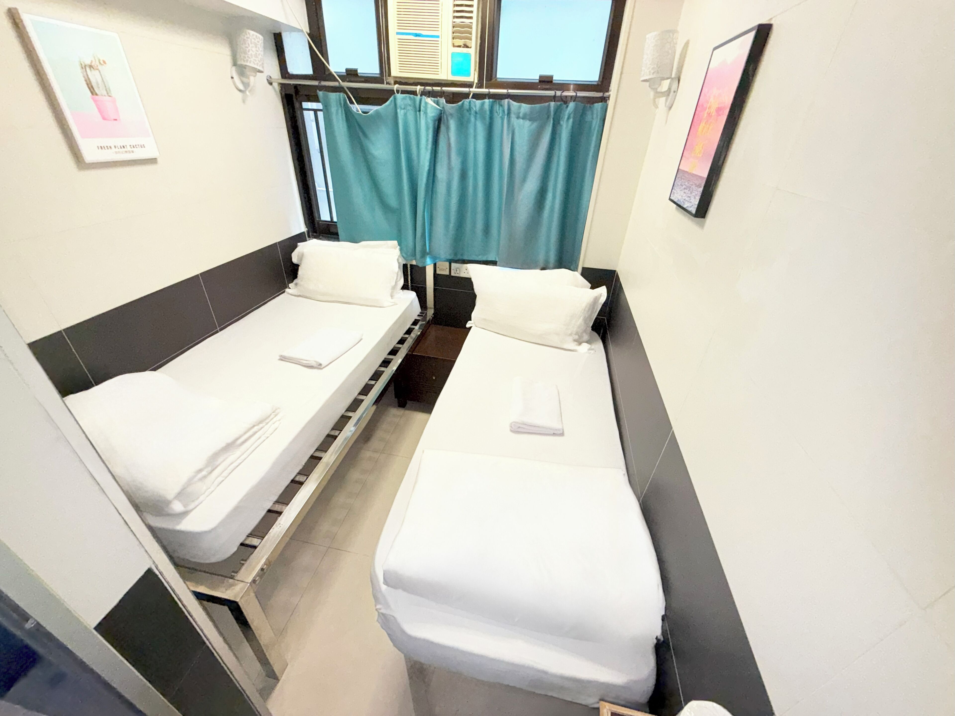 A small, clean room with two single beds, a window with curtains, and a wall-mounted light fixture.