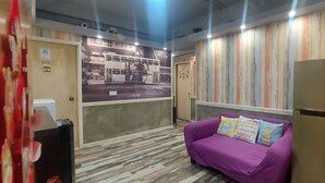 A room with a purple sofa, a refrigerator, a wall-mounted TV displaying a bus, and a wooden panel wall. - Apple Hostel (Kowloon)