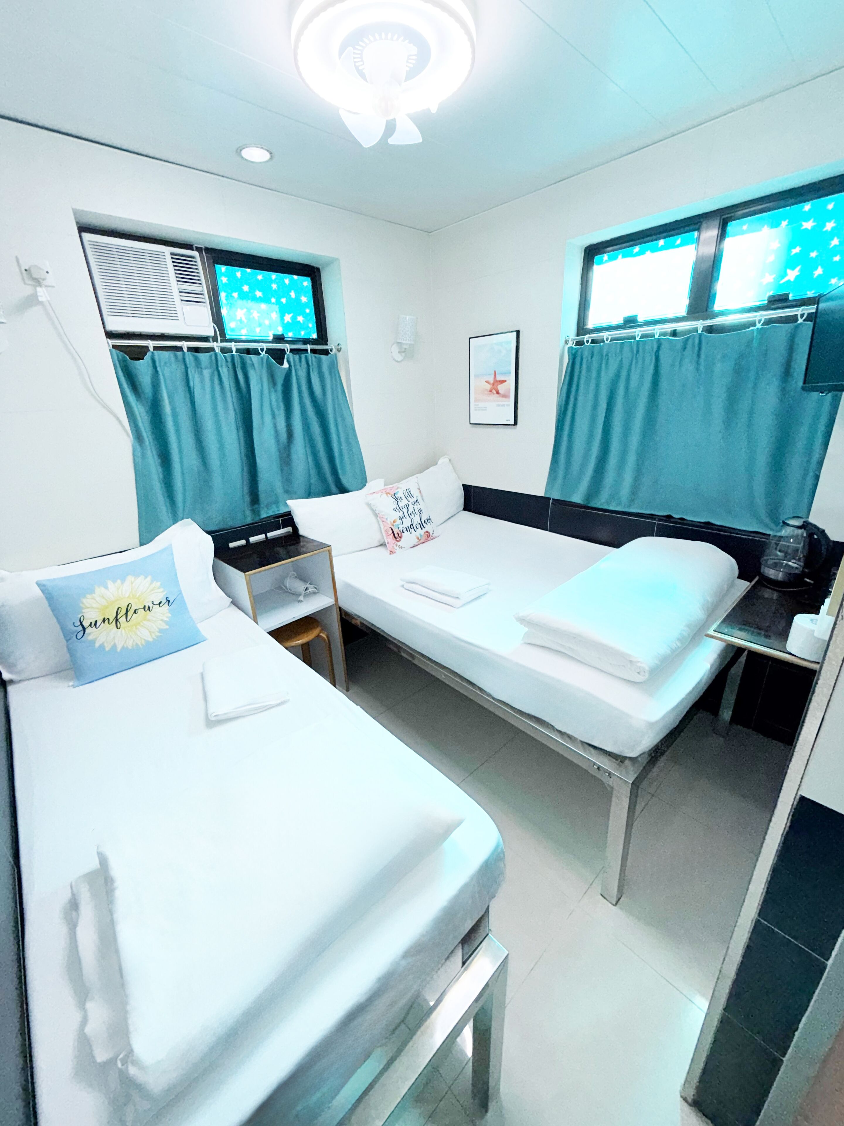 Quarto de hotel com duas camas de solteiro, uma mesa pequena, uma cadeira, escrivaninha e janela com cortinas.