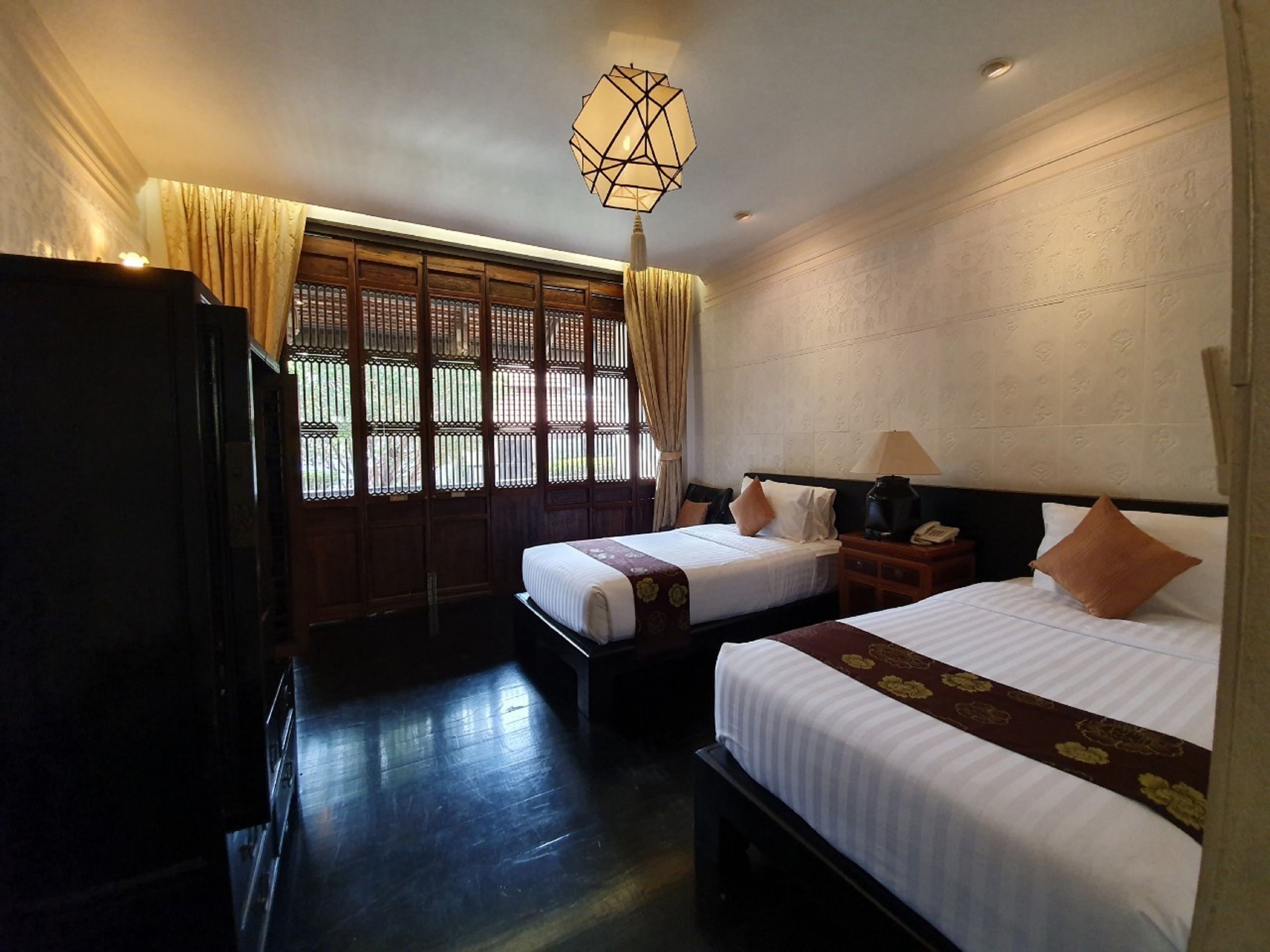 A hotel room with two beds, a wooden wardrobe, a desk with a lamp, and a window with curtains.