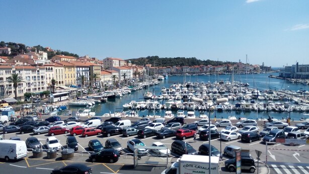 A marina with numerous boats docked, surrounded by parked cars and buildings with red-tiled roofs. - Hotel Résidence BEAR (Port-Vendres)