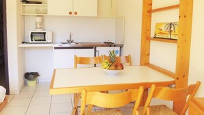A compact kitchen with white cabinets, a microwave, a sink, and a small stove. There is a dining table with chairs and a bowl of fruit on it. - Hotel Résidence BEAR (Port-Vendres)