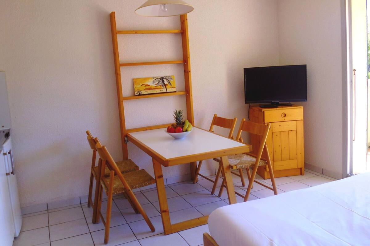 A room with a bed, a table with chairs, a television on a wooden cabinet, and a shelf with a painting.