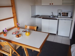 A compact kitchenette with a microwave, coffee maker, and sink. A breakfast table with croissants, a jug of orange juice, and a small potted plant is set. - Hotel Résidence BEAR (Port-Vendres)