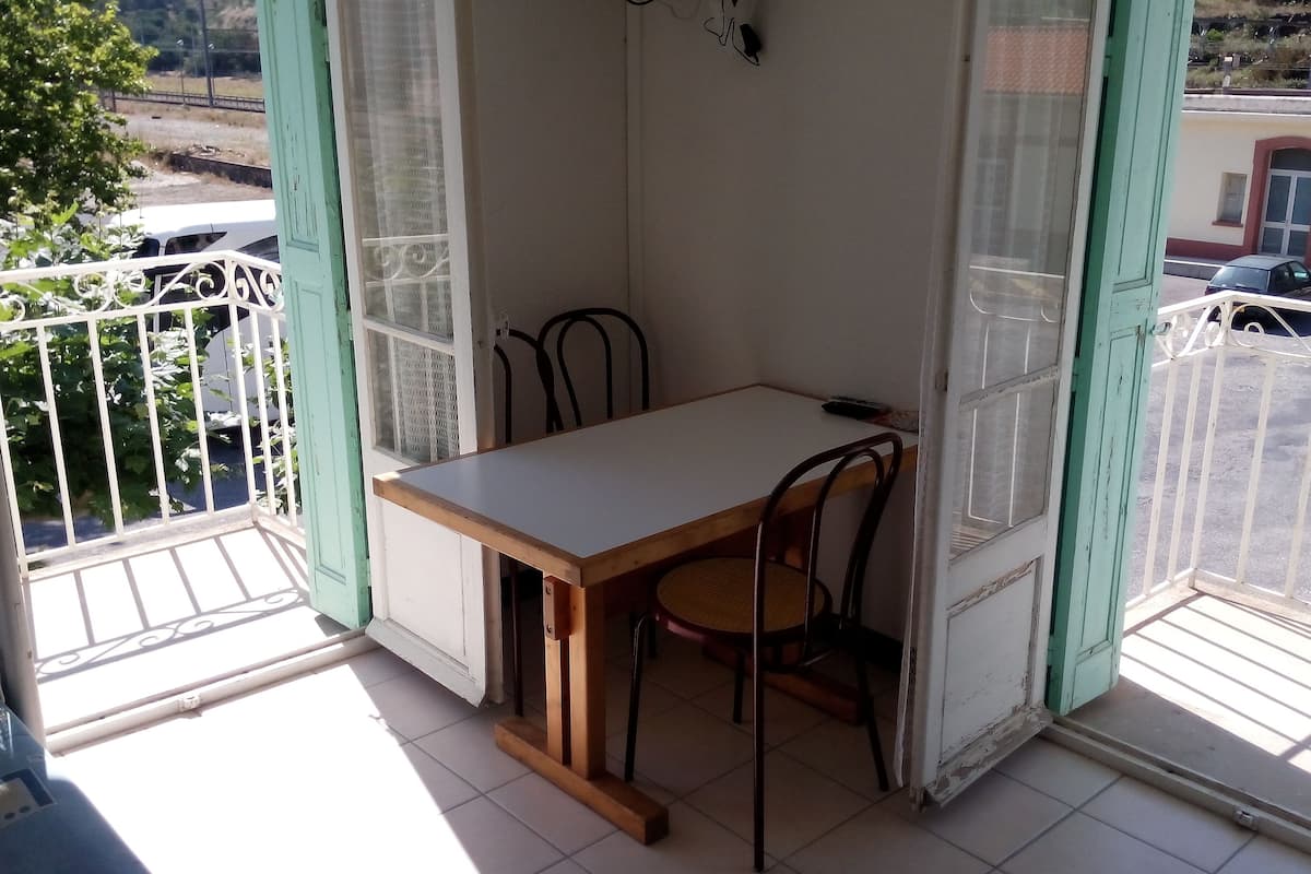 A small balcony with a table and two chairs, a view of the street, and a building in the background.