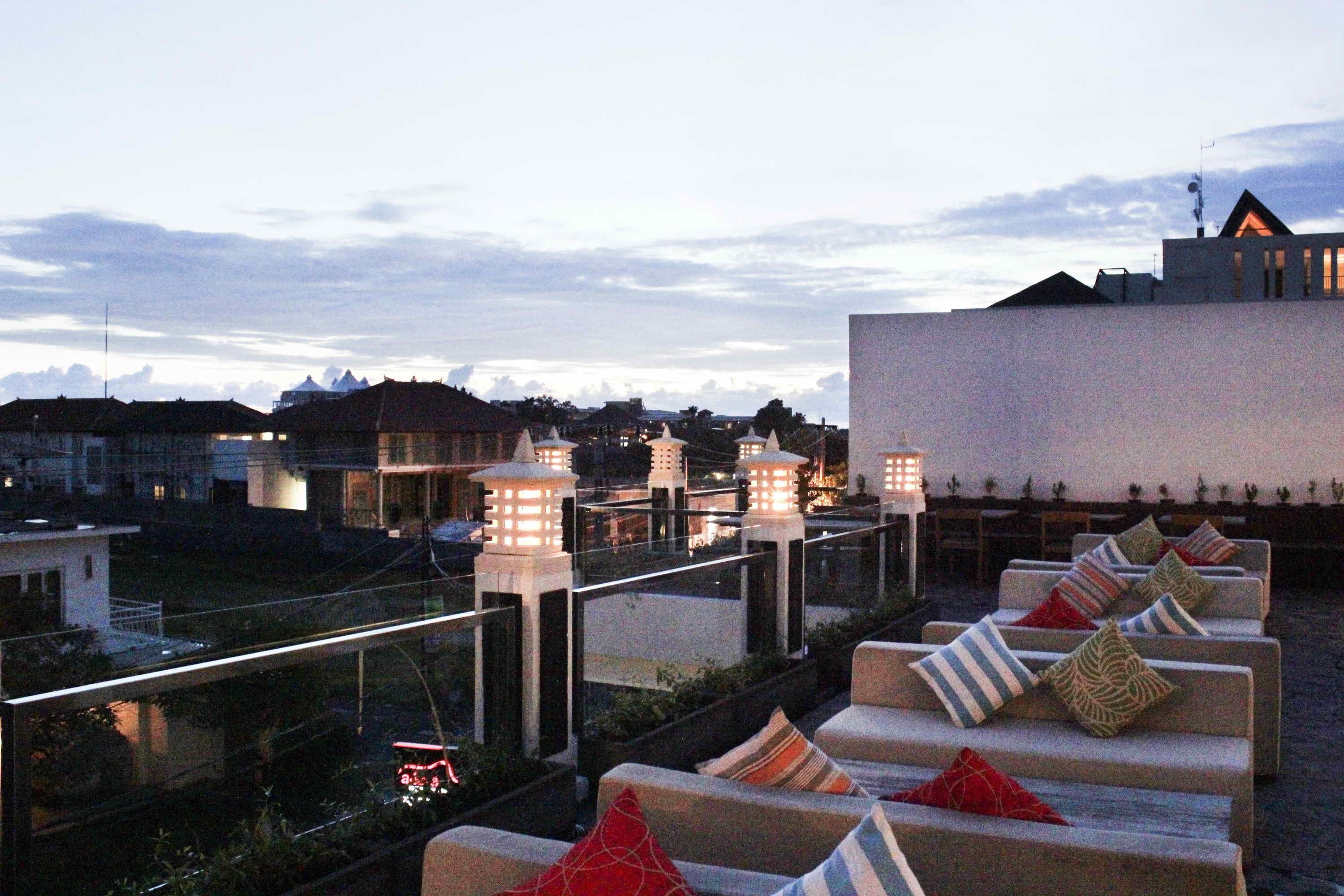 A rooftop terrace with outdoor seating and lanterns, overlooking a cityscape at dusk.