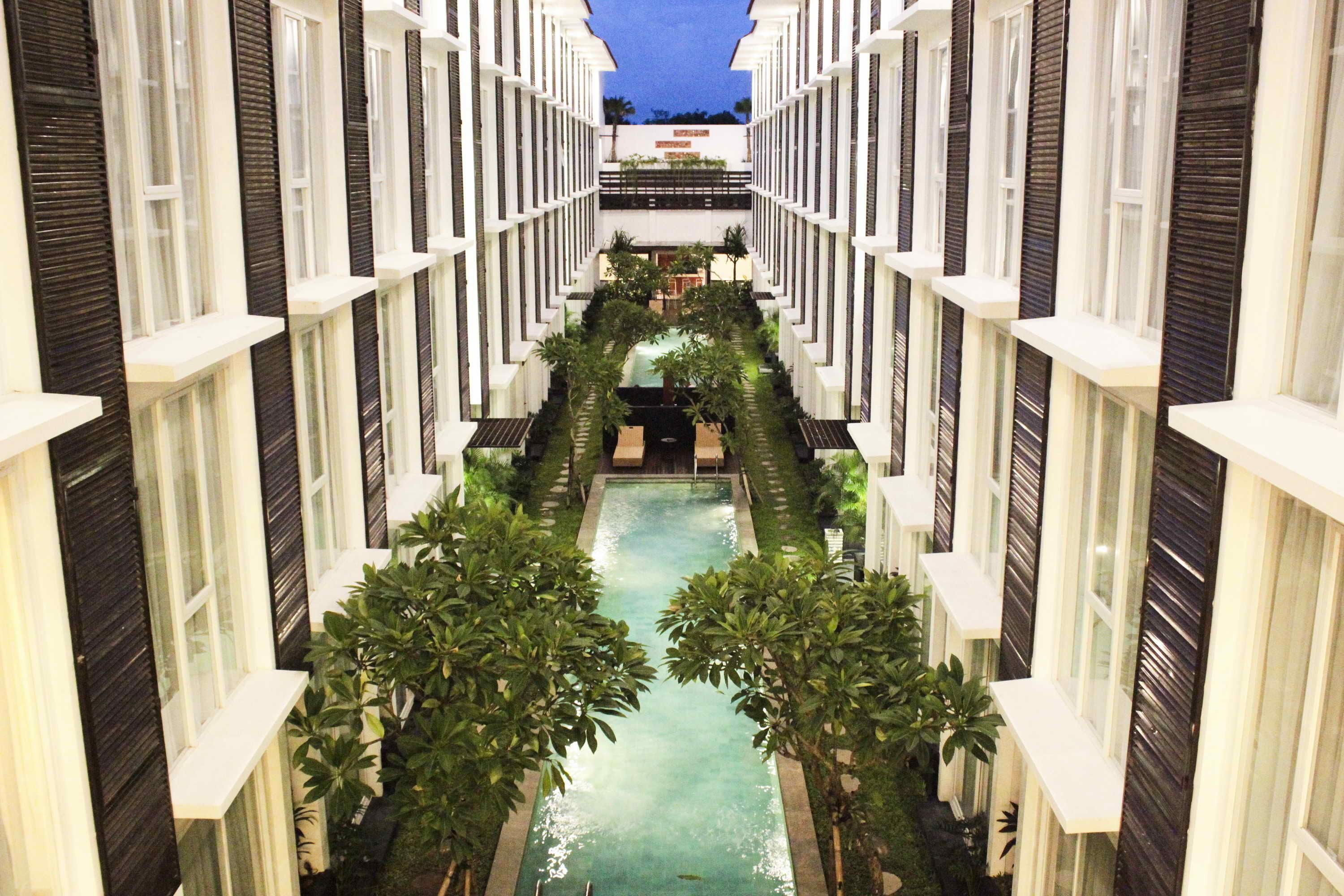 Corredor de hotel com piscina central, ladeado por árvores e janelas emolduradas em branco.