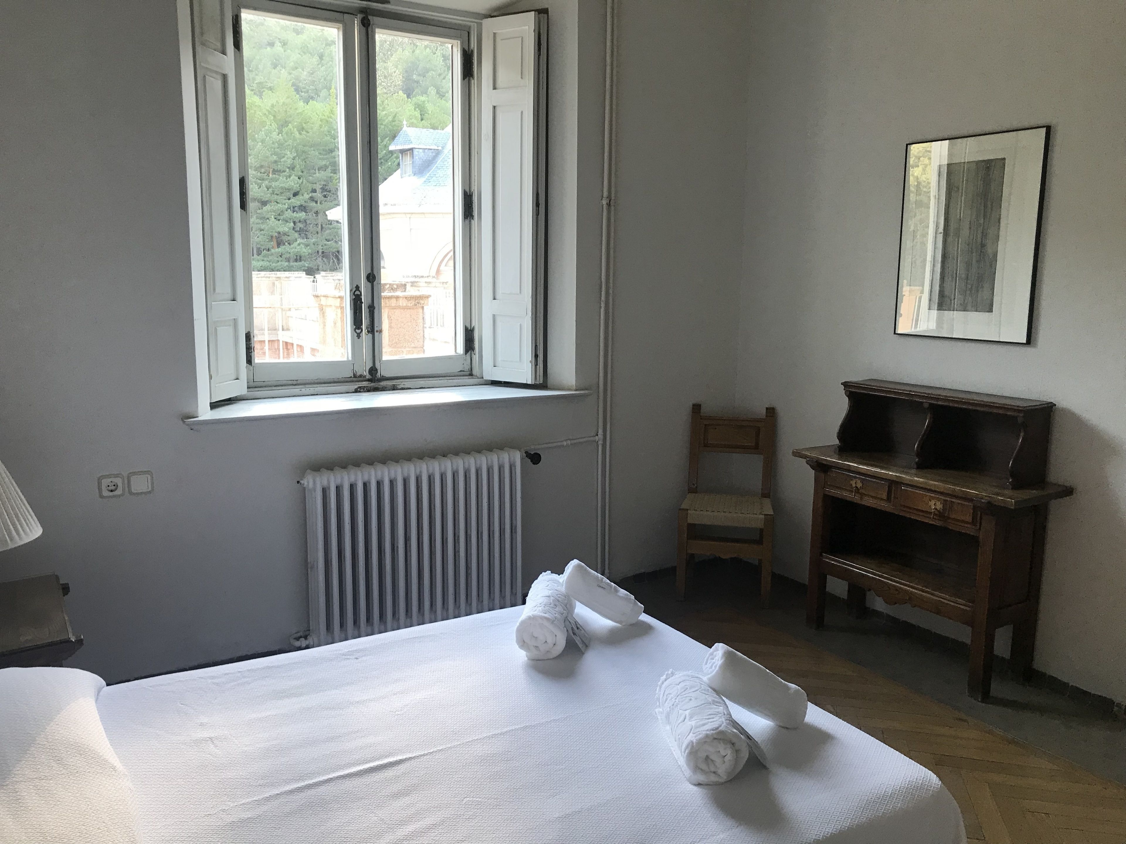 A room with a bed, a radiator, a chair, a desk, and a window with an open shutter.