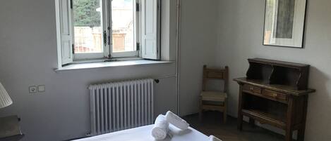 Una habitación con cama, radiador, silla, escritorio y ventana con persiana abierta.
