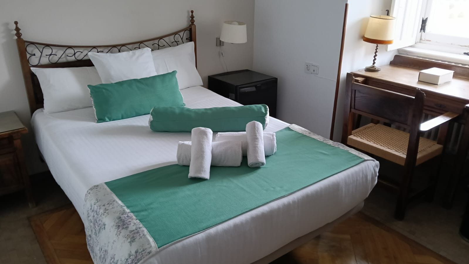 Un lit bien fait, avec une literie blanche et verte, une petite table de chevet avec une lampe, et une chaise en bois avec un siège en osier.