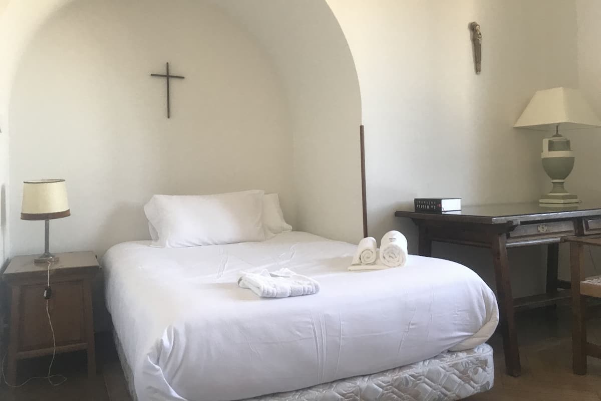 A neatly made bed with white linens, a bedside lamp, a wooden nightstand, and a desk with a lamp.