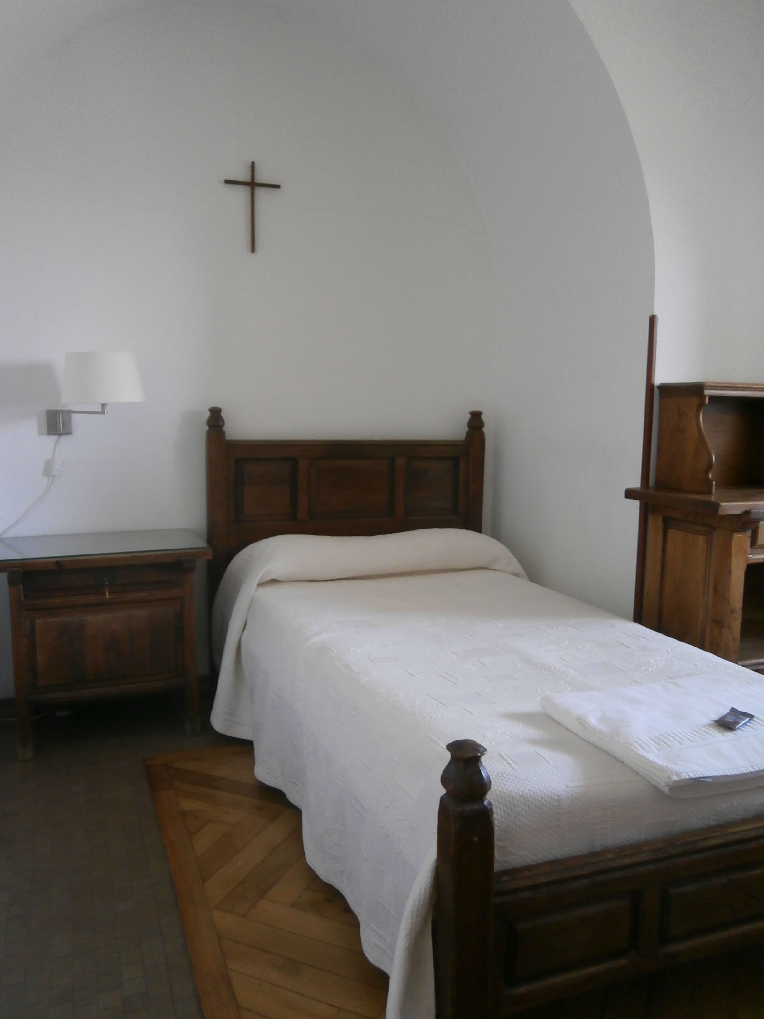 Un dormitorio con una cama de madera, una mesita de noche, una lámpara y una cruz en la pared.