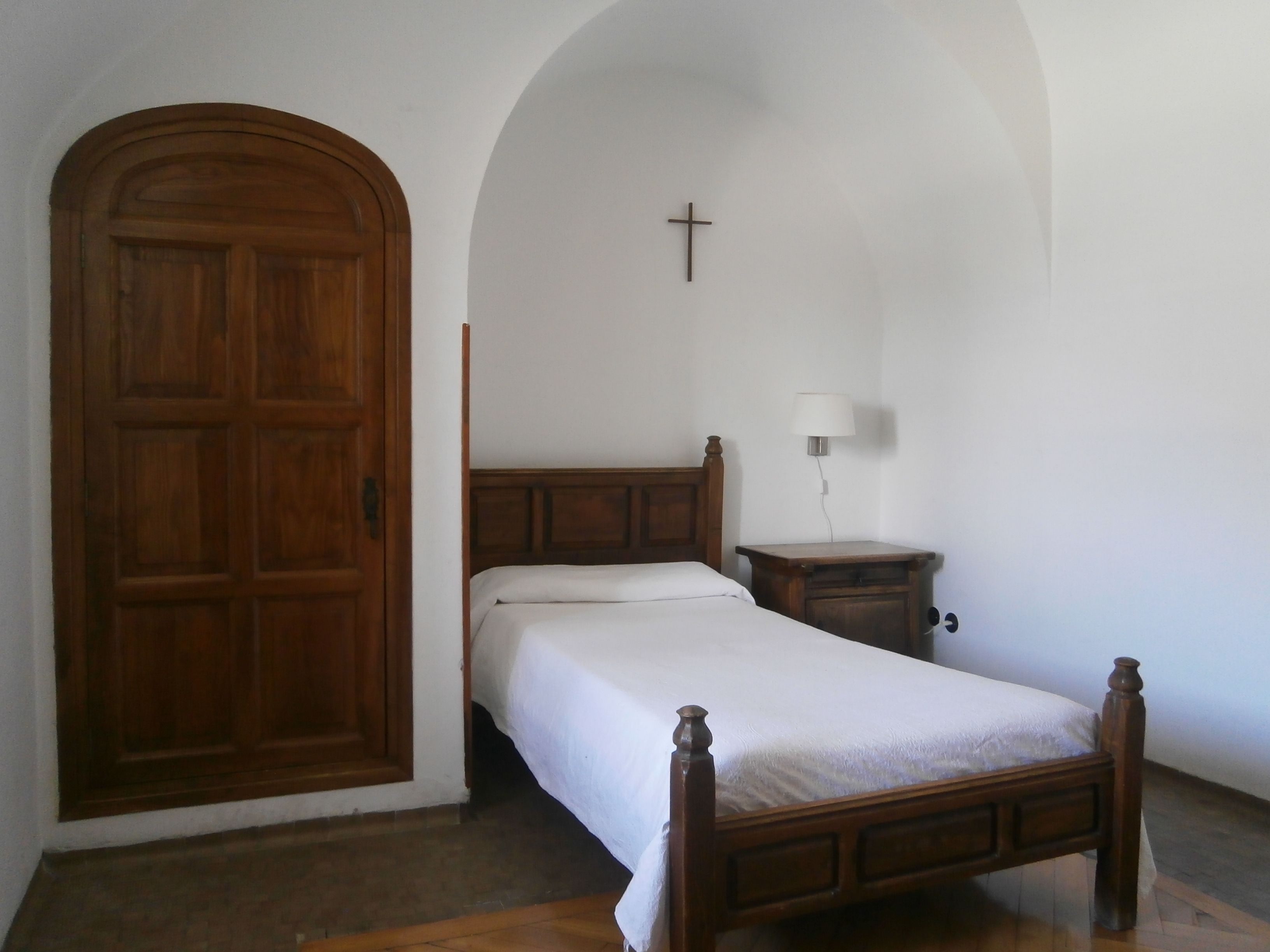 Un dormitorio con puerta de madera, cama con cabecero de madera, mesita de noche con lámpara y una cruz en la pared.