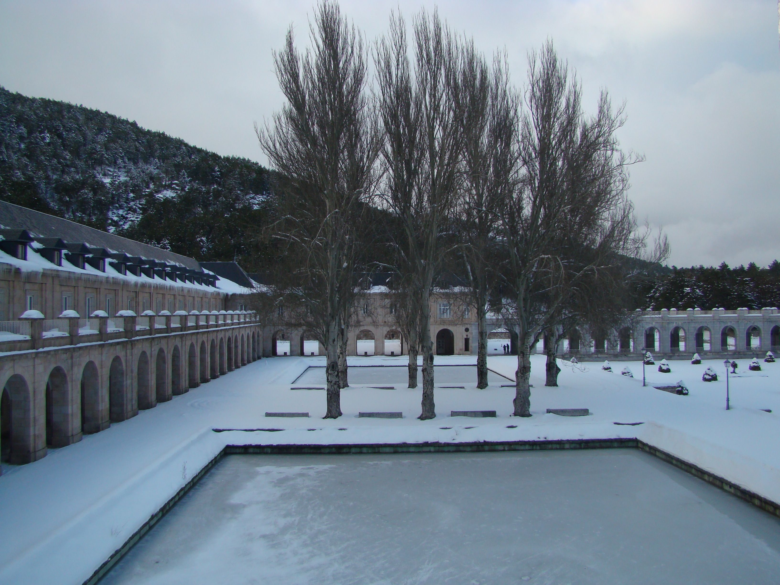 Un patio con suelo cubierto de nieve, una piscina y un edificio con ventanas arqueadas y un balcón.