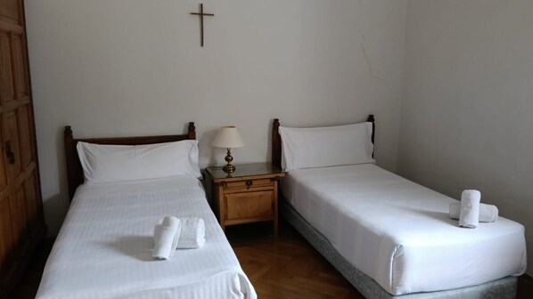 Una habitación con dos camas individuales, cada una con cabecero de madera, ropa de cama blanca y una toalla doblada sobre la cama. Hay una mesita de noche de madera con una lámpara entre las camas.