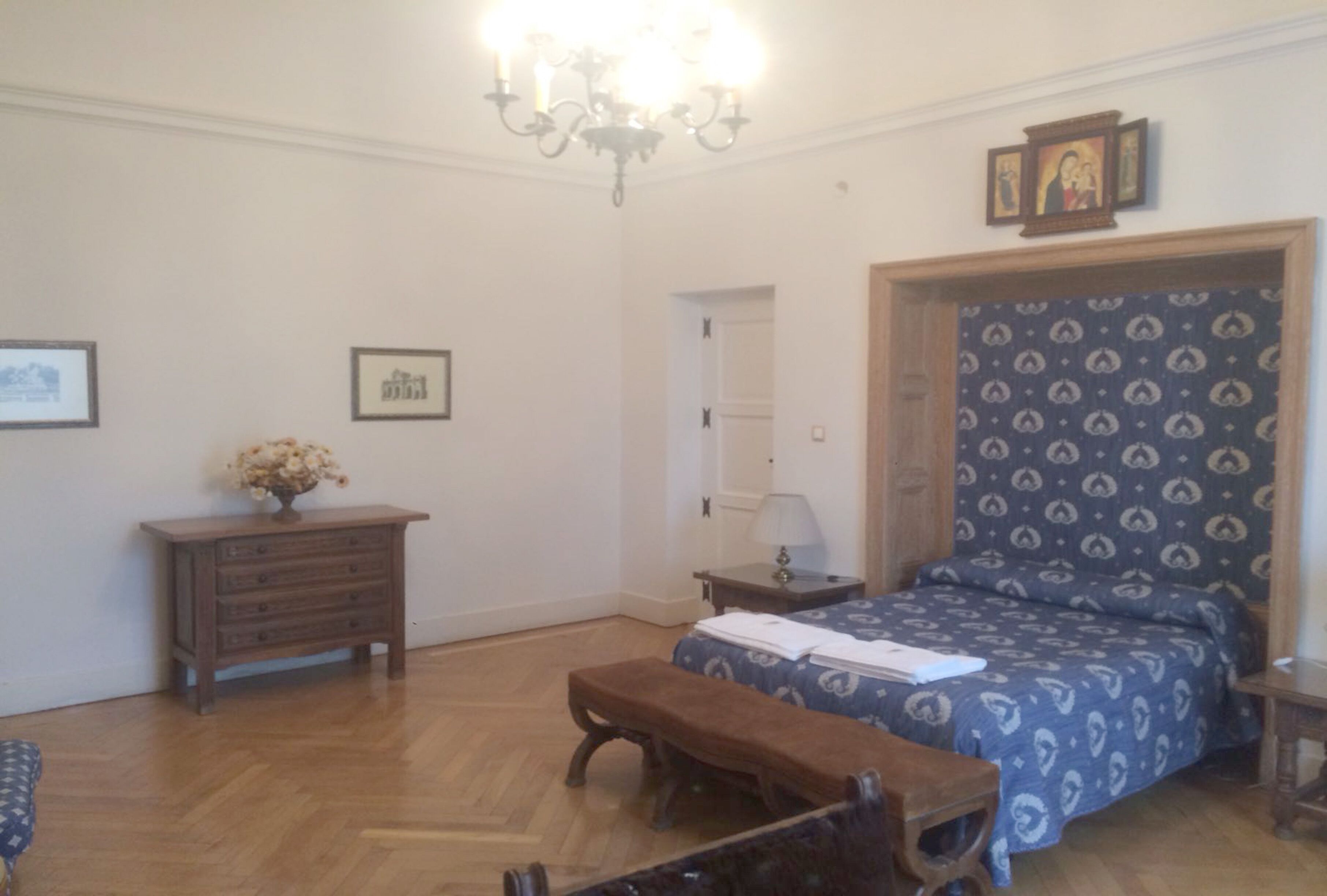 A bedroom with a wooden floor, a bed with a patterned headboard, a bench, a dresser, and a chandelier.