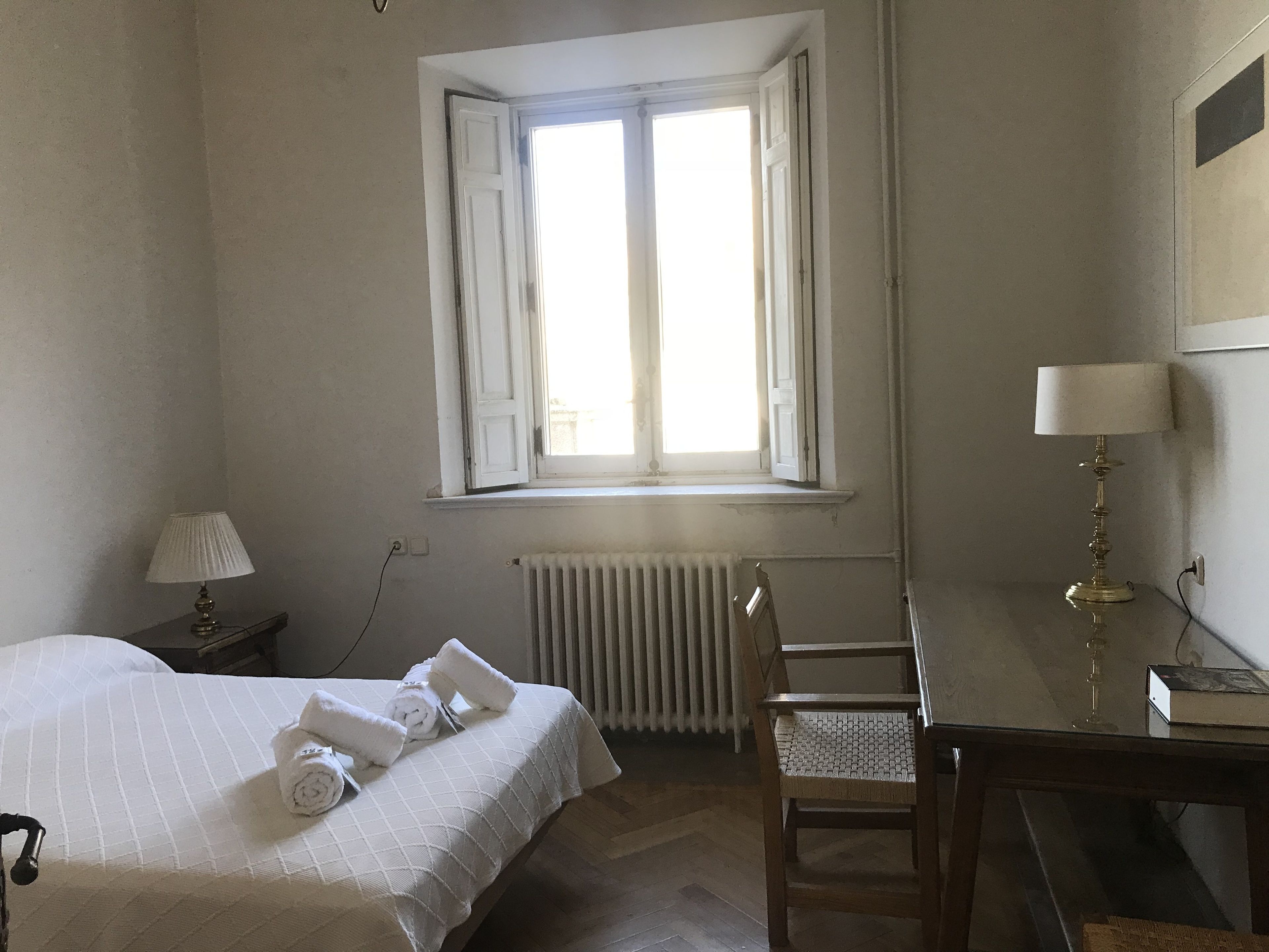 Habitación de hotel con cama, escritorio, silla, radiador y ventana con cortinas.