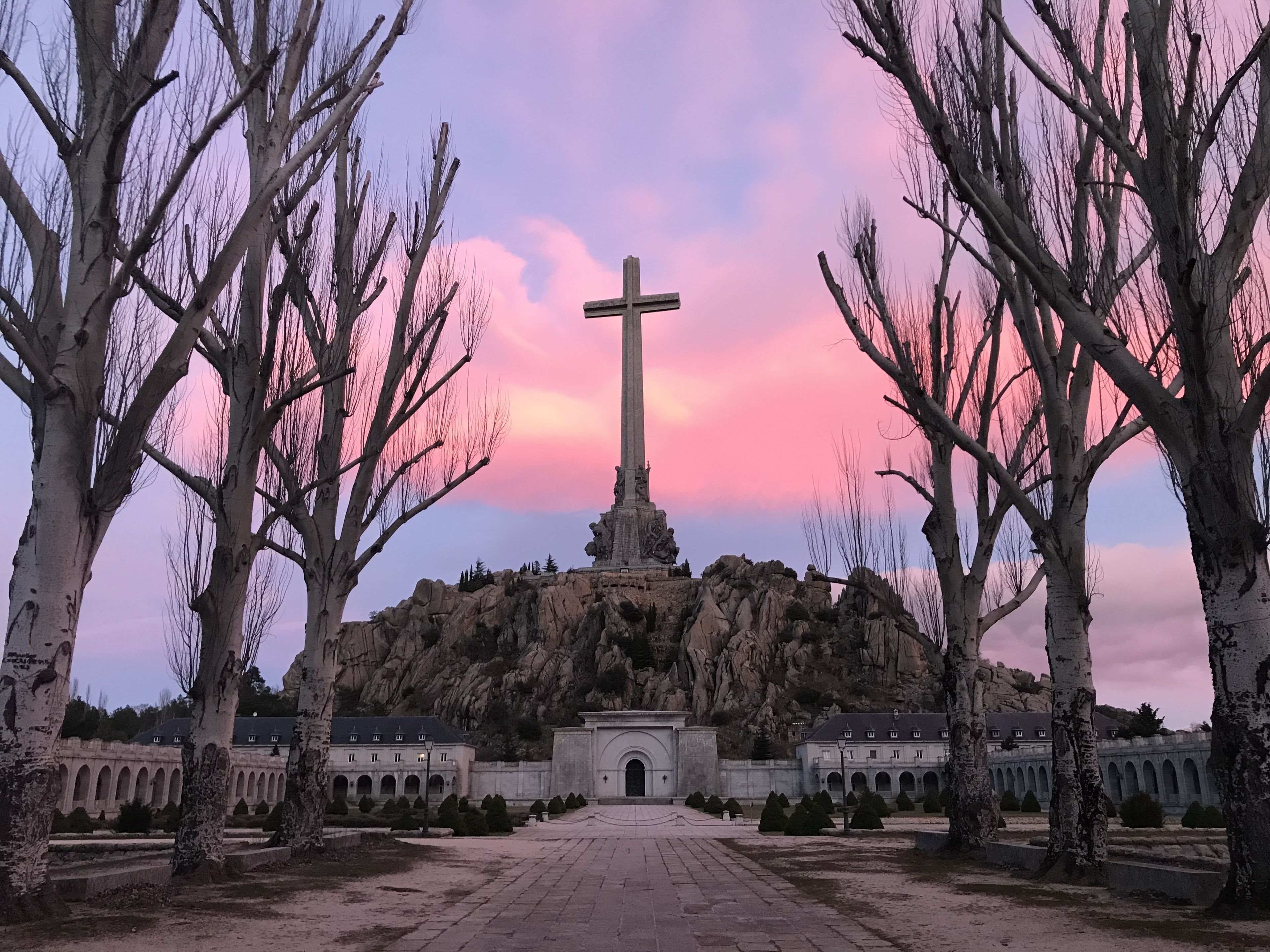 Un monumento con una gran cruz rodeado de árboles desnudos y un edificio con múltiples arcos al fondo.