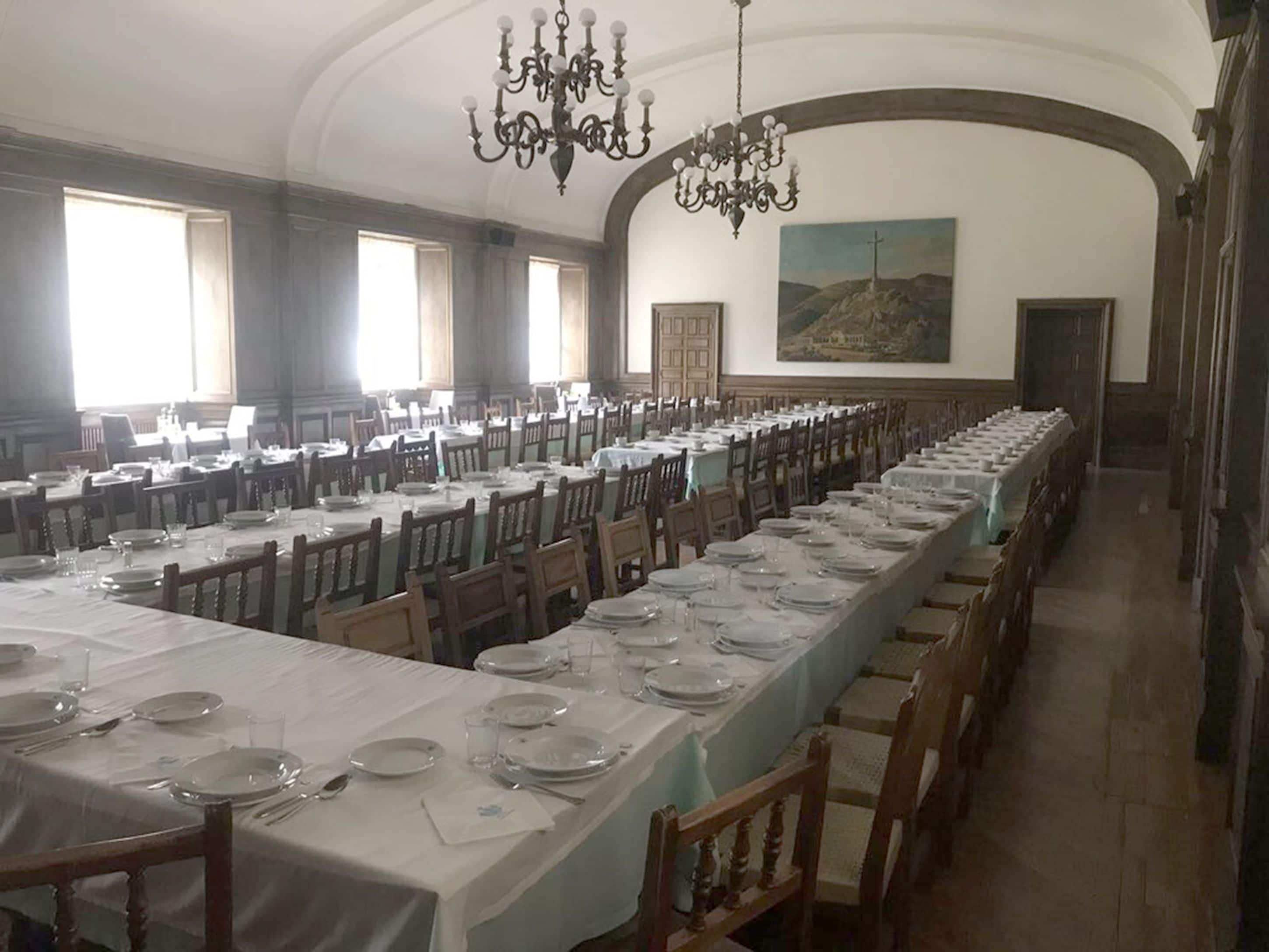 A long dining hall with multiple tables set for a meal, large windows, and a chandelier.