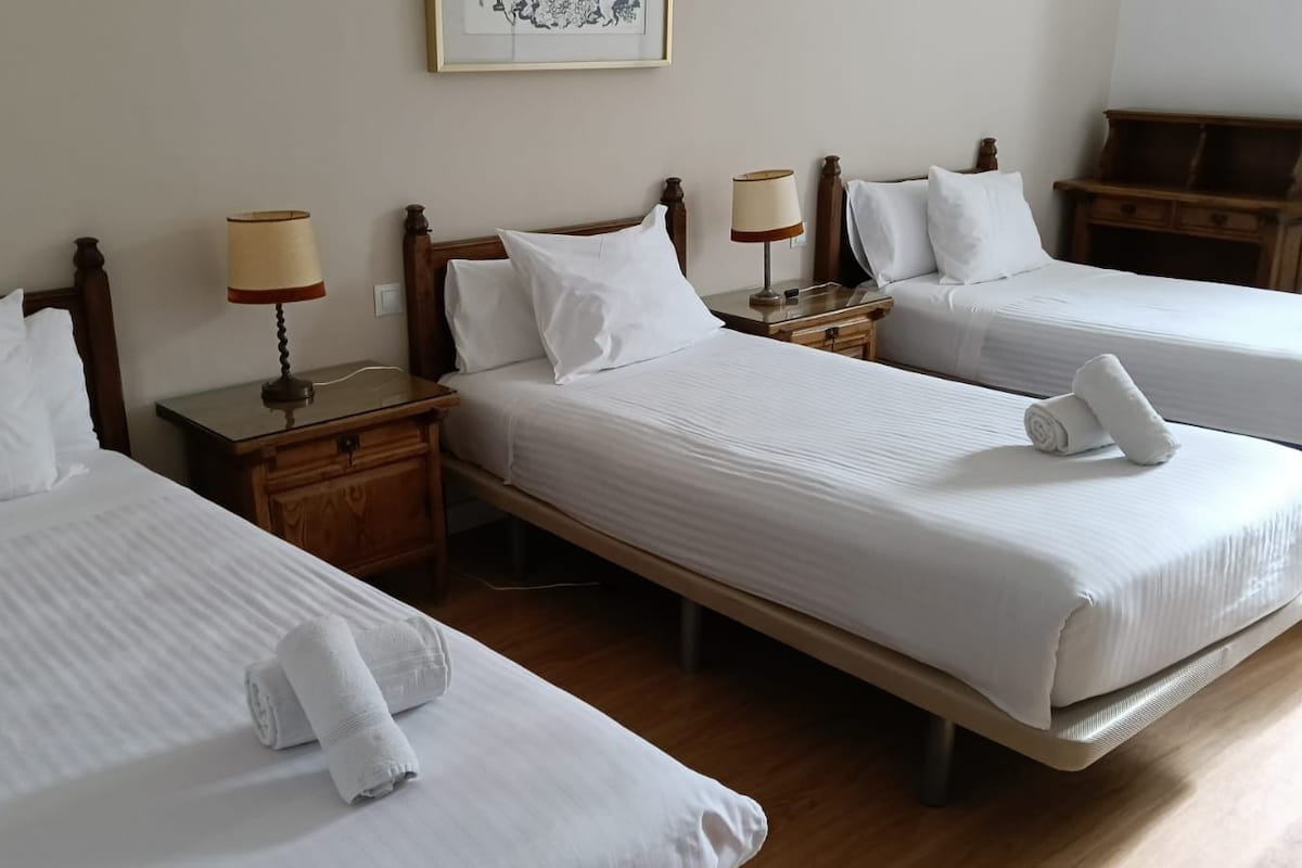 A hotel room with two single beds, each with a wooden headboard, white linens, and a rolled towel on the bed. There are wooden nightstands with lamps and a small wooden table between the beds.
