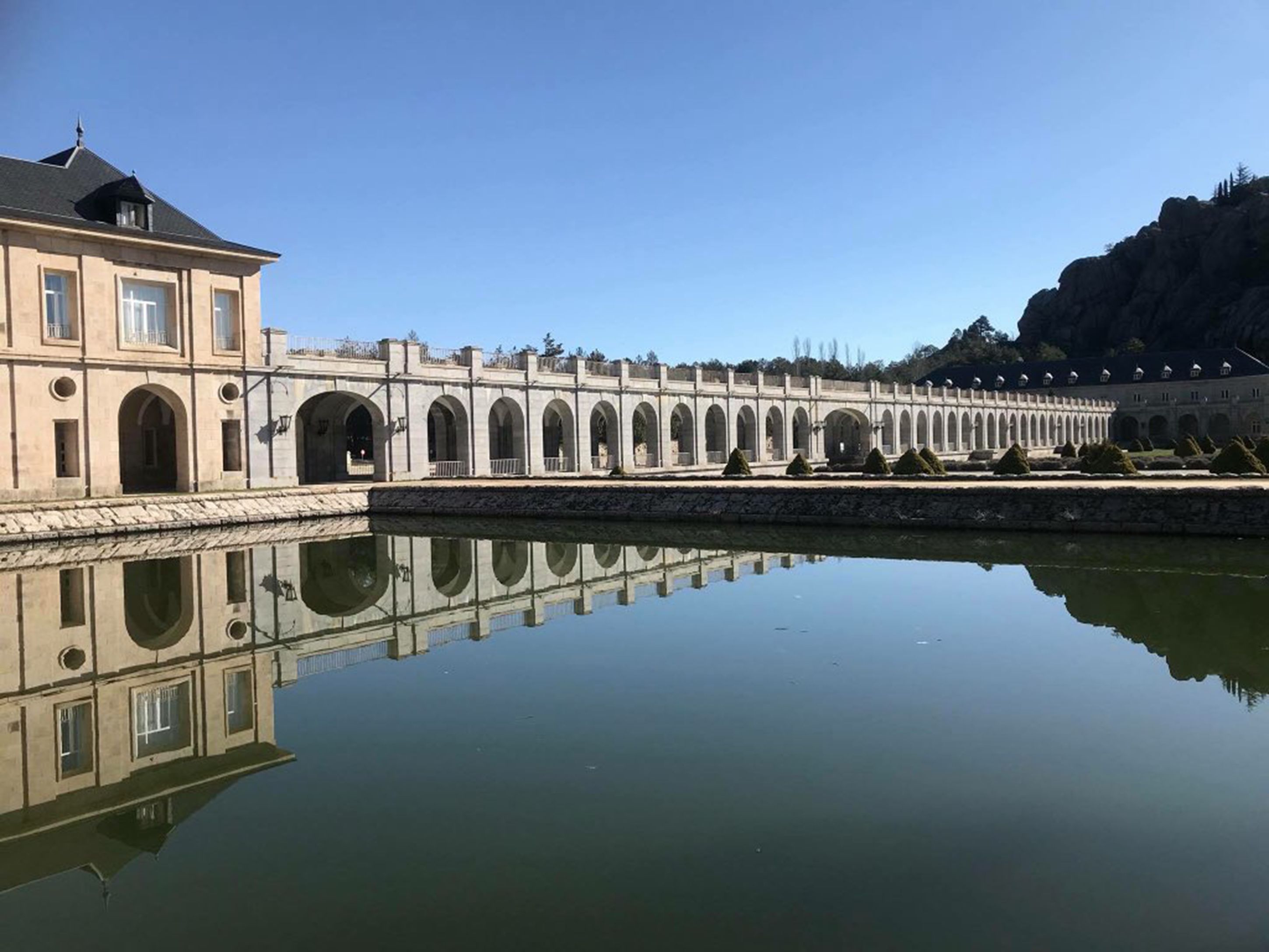 A long, arched bridge over a calm body of water, reflecting the structure and surrounding greenery.