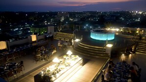 Un área de comedor en la azotea con varias mesas y sillas, un elemento decorativo central de iluminación y vistas de la ciudad al atardecer.