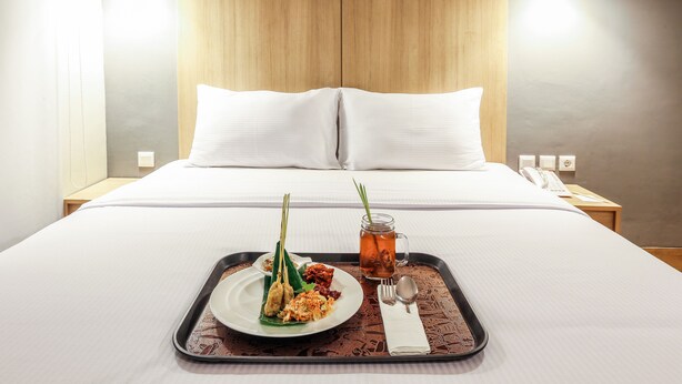 A neatly made bed with white linens, a tray with a plate of food and a drink, and a bedside table with a lamp and a phone. - Hotel Citradream Semarang (Semarang)