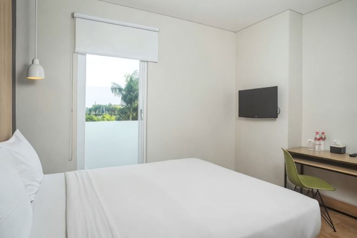 Habitación de hotel con cama, escritorio, silla, televisor y puerta que da a un balcón con vistas a zonas verdes.