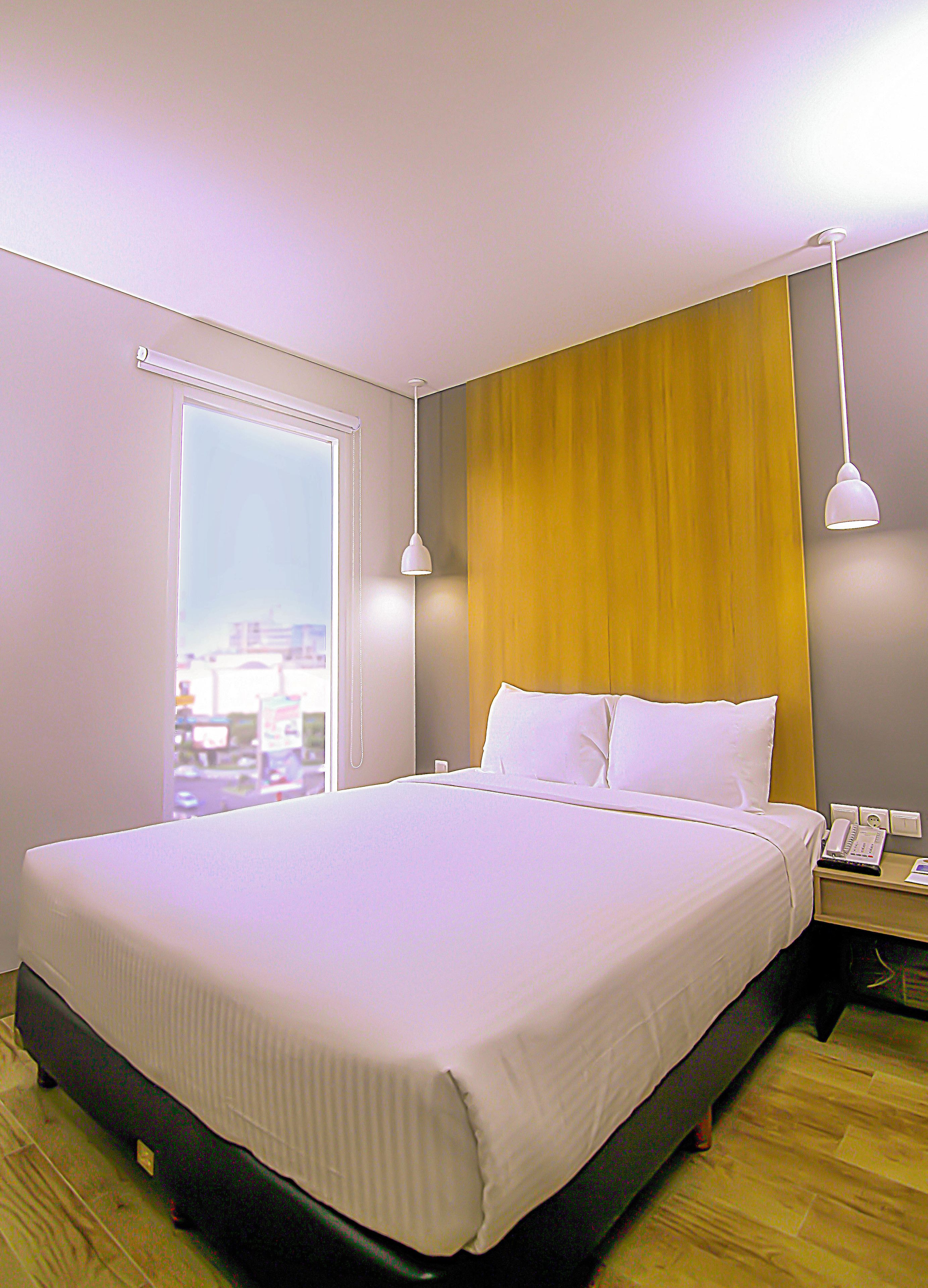 A hotel room with a large bed, a wooden headboard, a bedside table with a lamp, and a window with a city view.