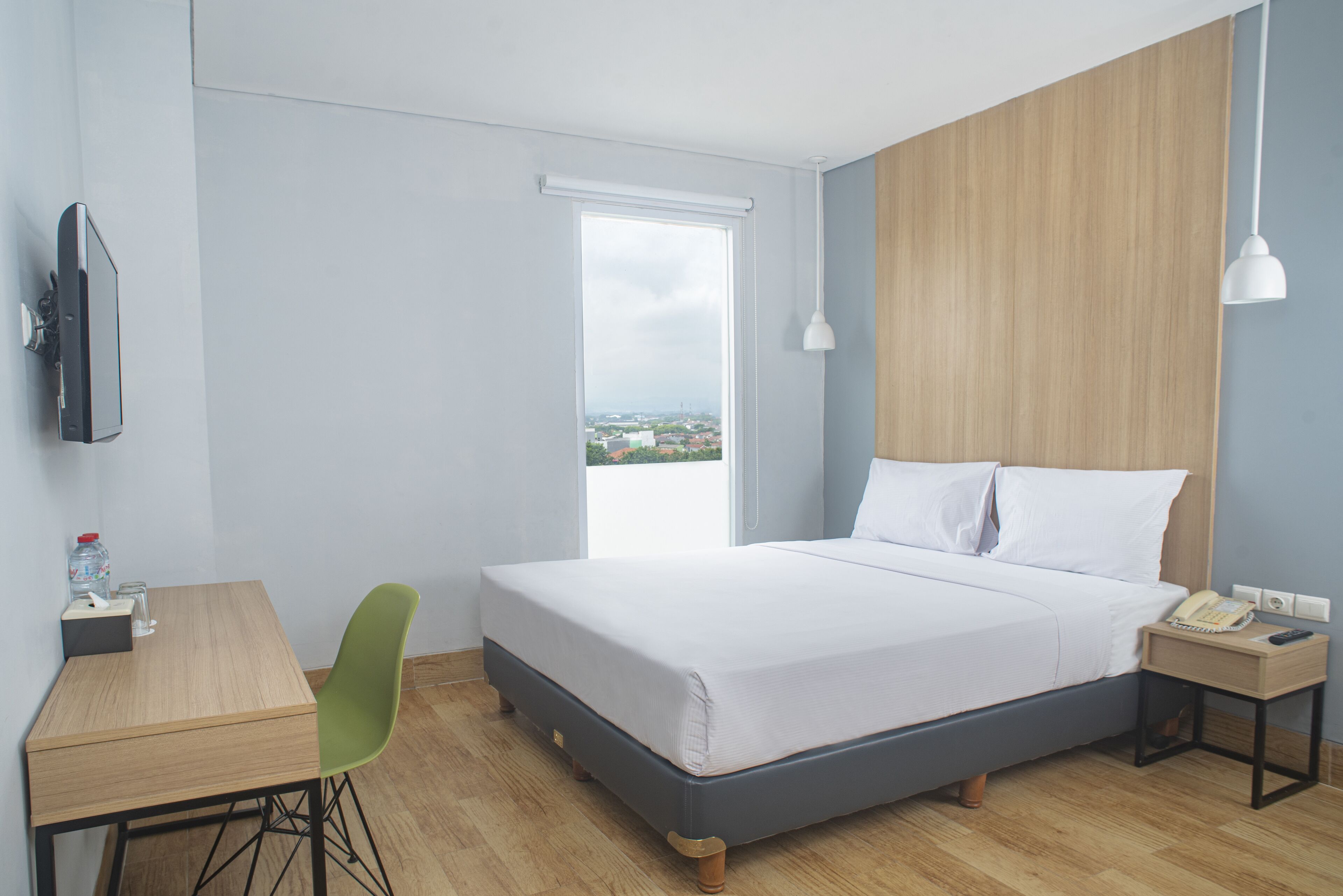 Habitación de hotel con cama, escritorio, silla, televisor, ventana con vistas y mesita de noche con teléfono y lámpara.
