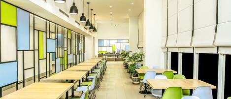 A modern cafeteria with long tables and chairs, a kitchen area in the background, and pendant lighting.