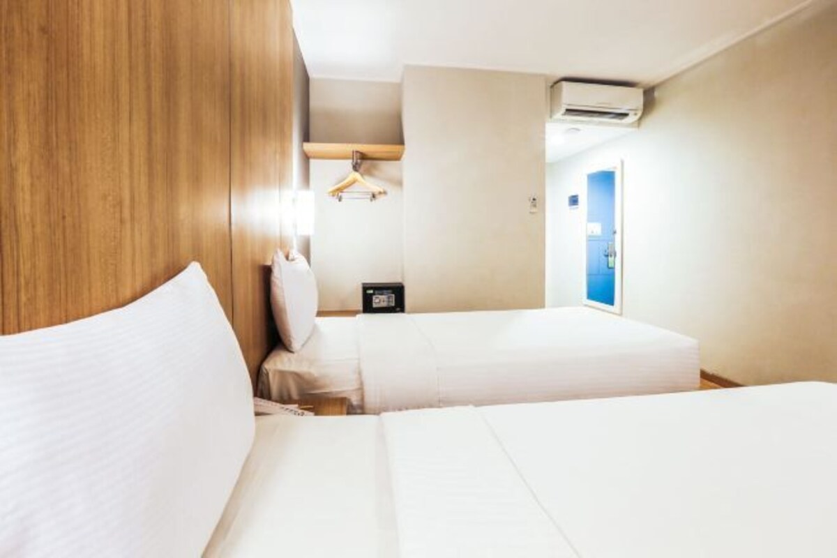Une chambre d’hôtel avec deux lits, une tête de lit en bois, une armoire, une table de chevet avec une horloge et un appareil de climatisation.