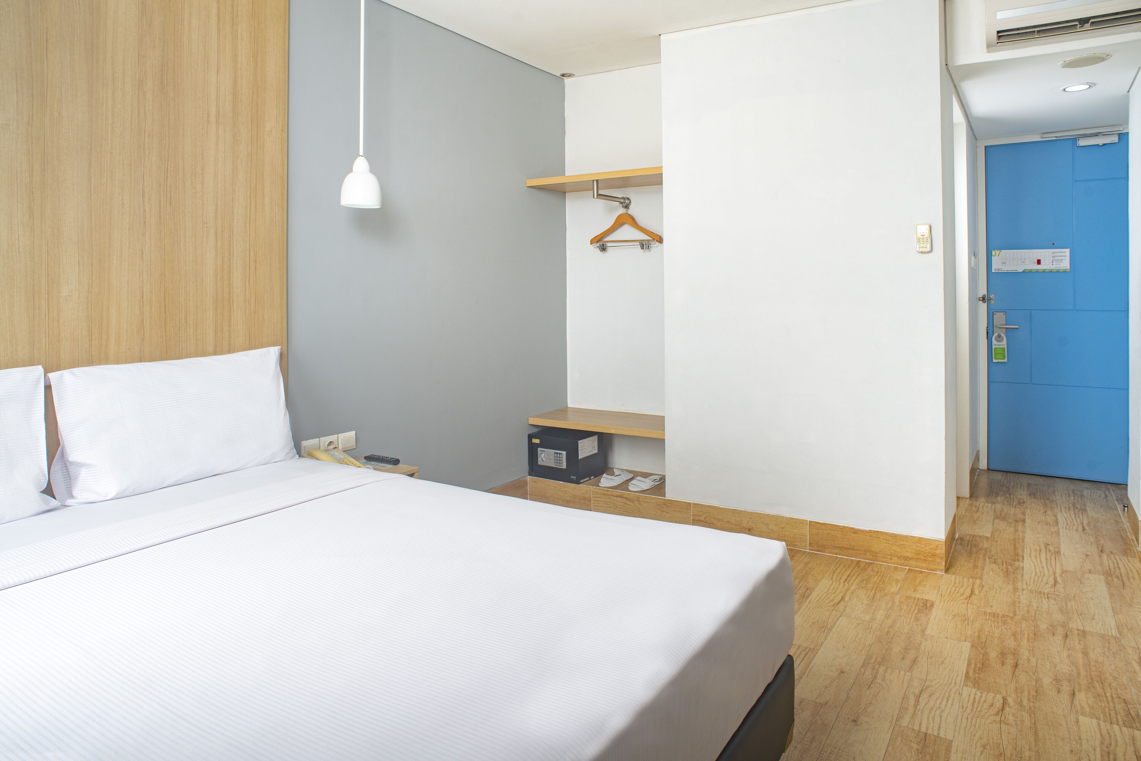 Habitació d'hotel amb llit, una taula de nit, una perxa per a abrics, una petita estant de fusta i una porta blava.