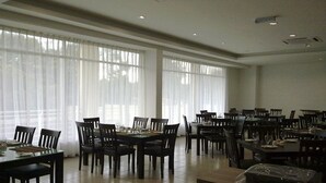 Un comedor amplio con varias mesas y sillas, ventanas grandes con cortinas ligeras y piso de baldosas.