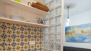 Cocina compacta con encimera de madera, pileta integrada y lavavajillas blanco. Se observa un sofá gris y un cuadro llamativo al fondo.