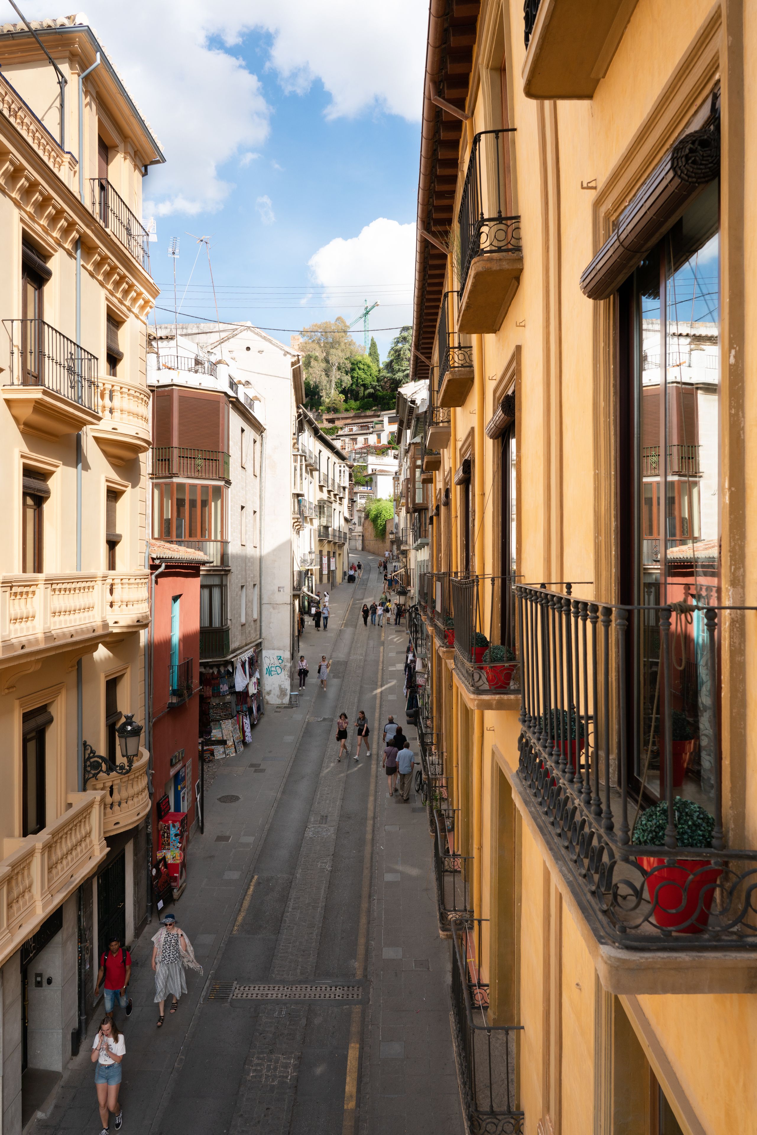 A narrow street lined with multi-story buildings, featuring balconies with potted plants and a clear sky above.