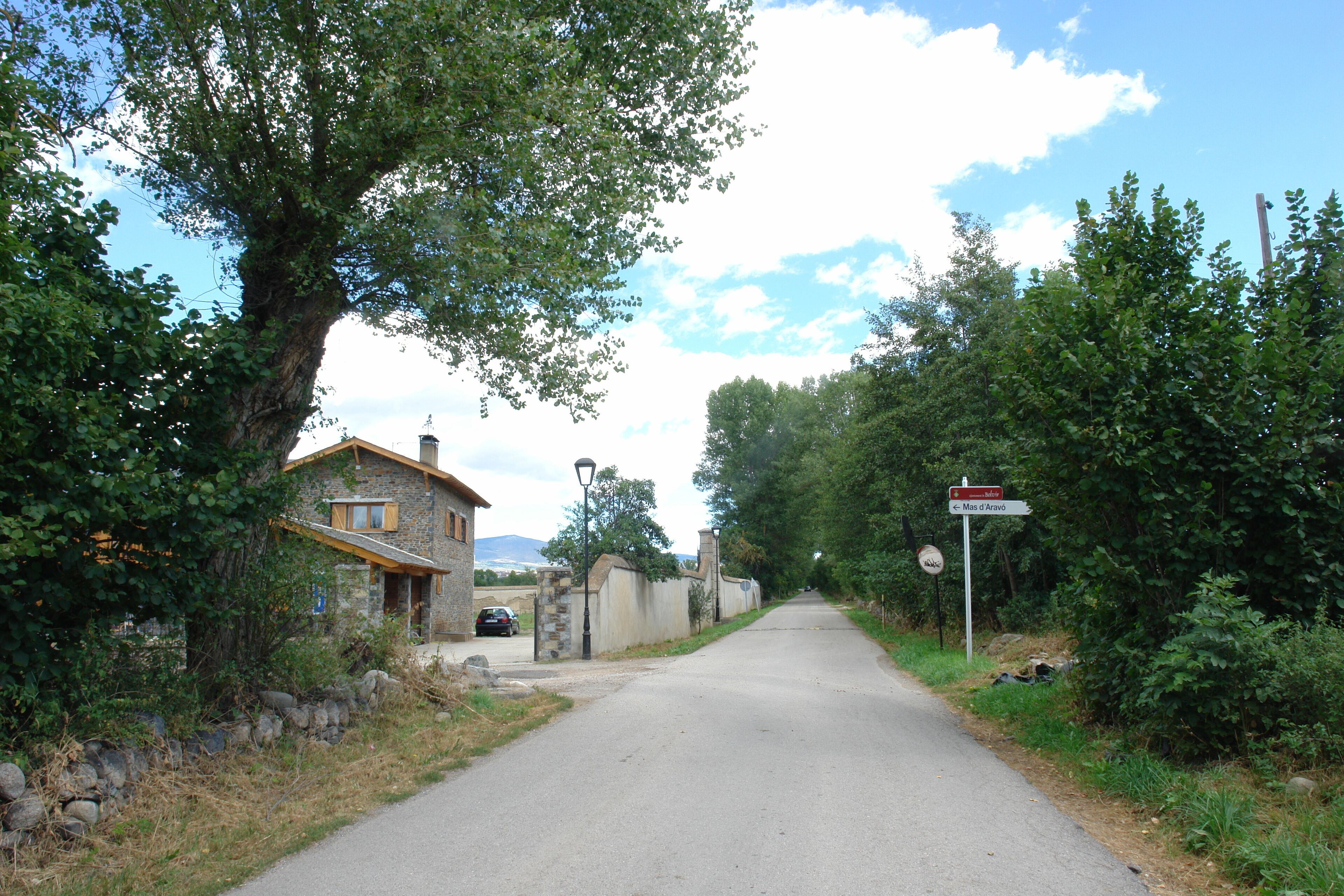 A narrow road flanked by stone walls and trees, leading to a stone house with a wooden roof.