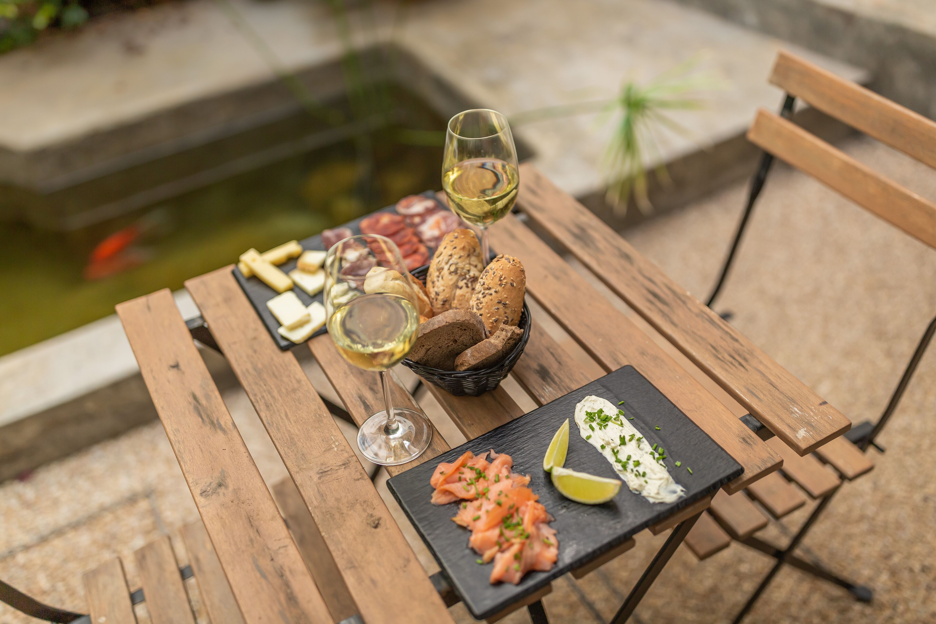 Une table en bois, sur laquelle on trouve une variété d'aliments, notamment du pain, du fromage et du saumon fumé, accompagnée d'un verre de vin blanc.