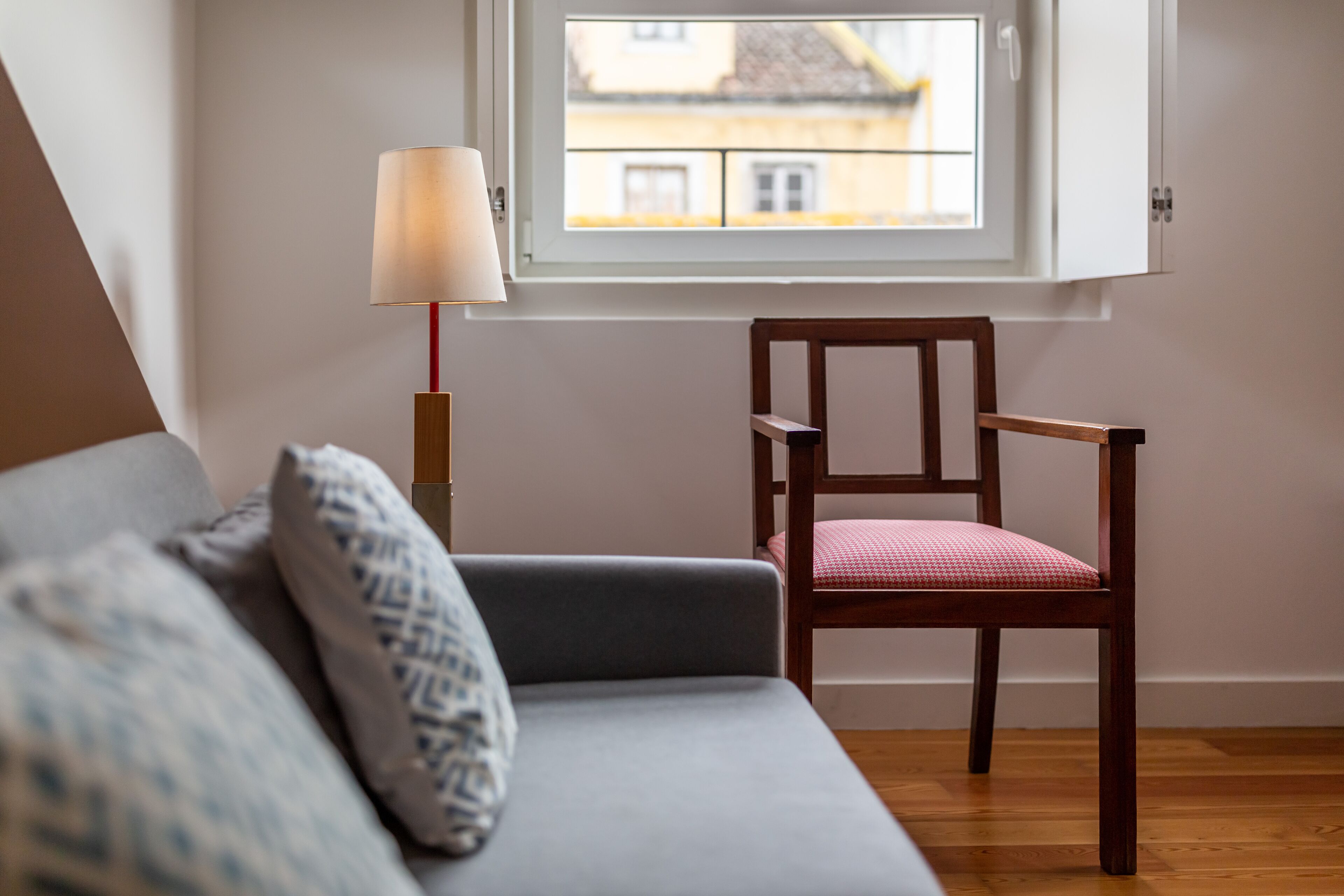 A room with a grey sofa, a wooden chair with a red cushion, a floor lamp, and a window overlooking a building.