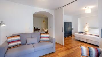 A modern living room with a grey sofa, colorful cushions, a wooden floor, and a dining area in the background.
