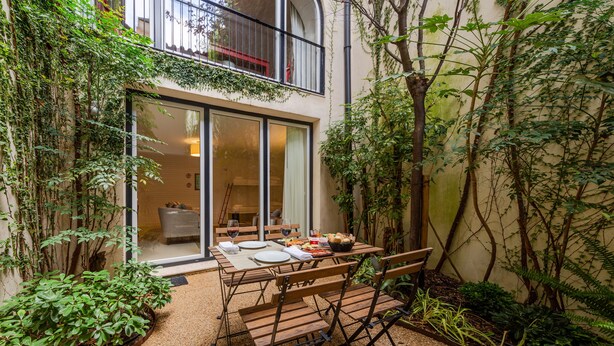 An outdoor dining area with a table and chairs, surrounded by plants and a building entrance.