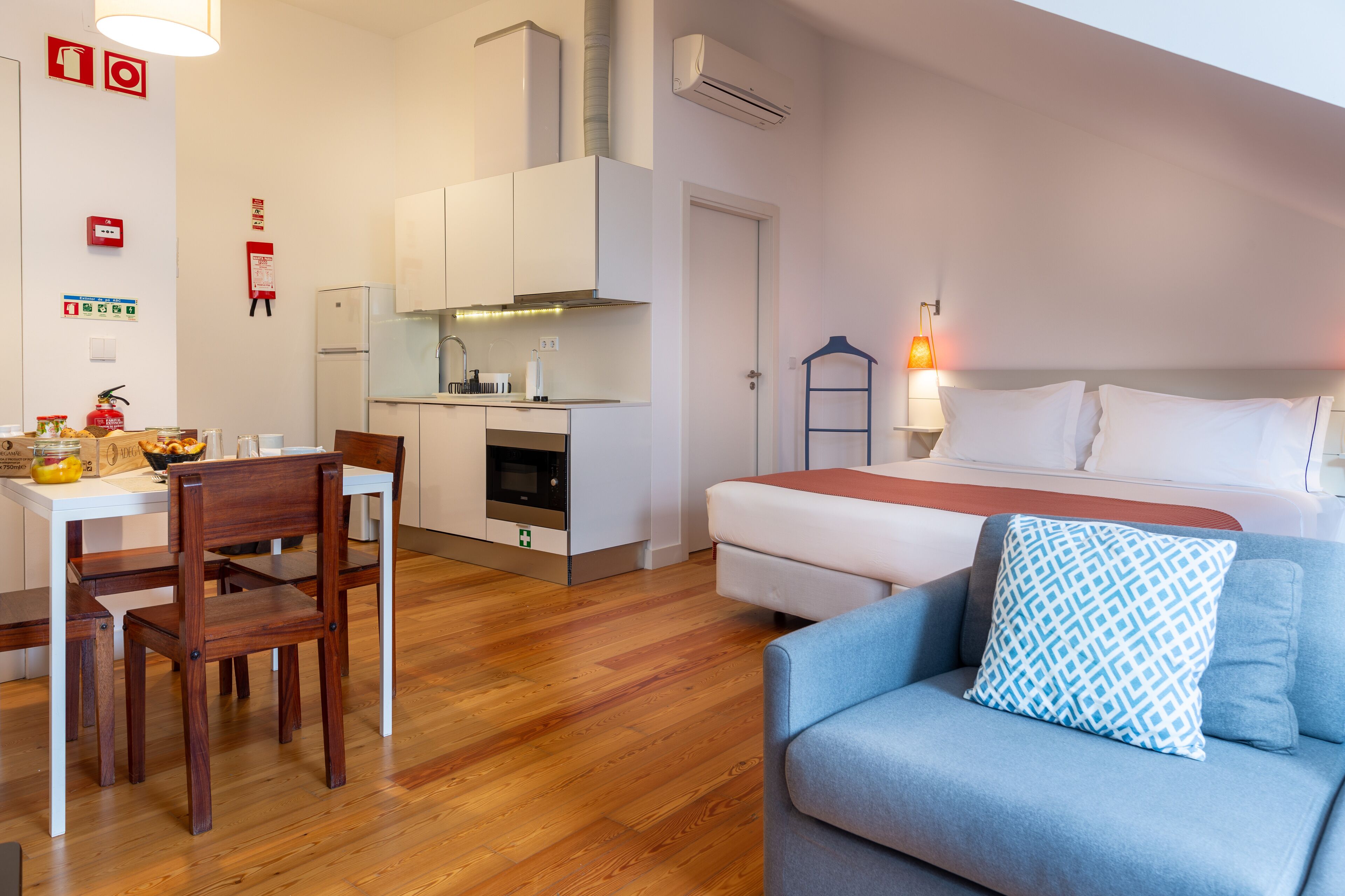 A compact hotel room with a kitchenette, a dining area, and a bed with a patterned pillow.