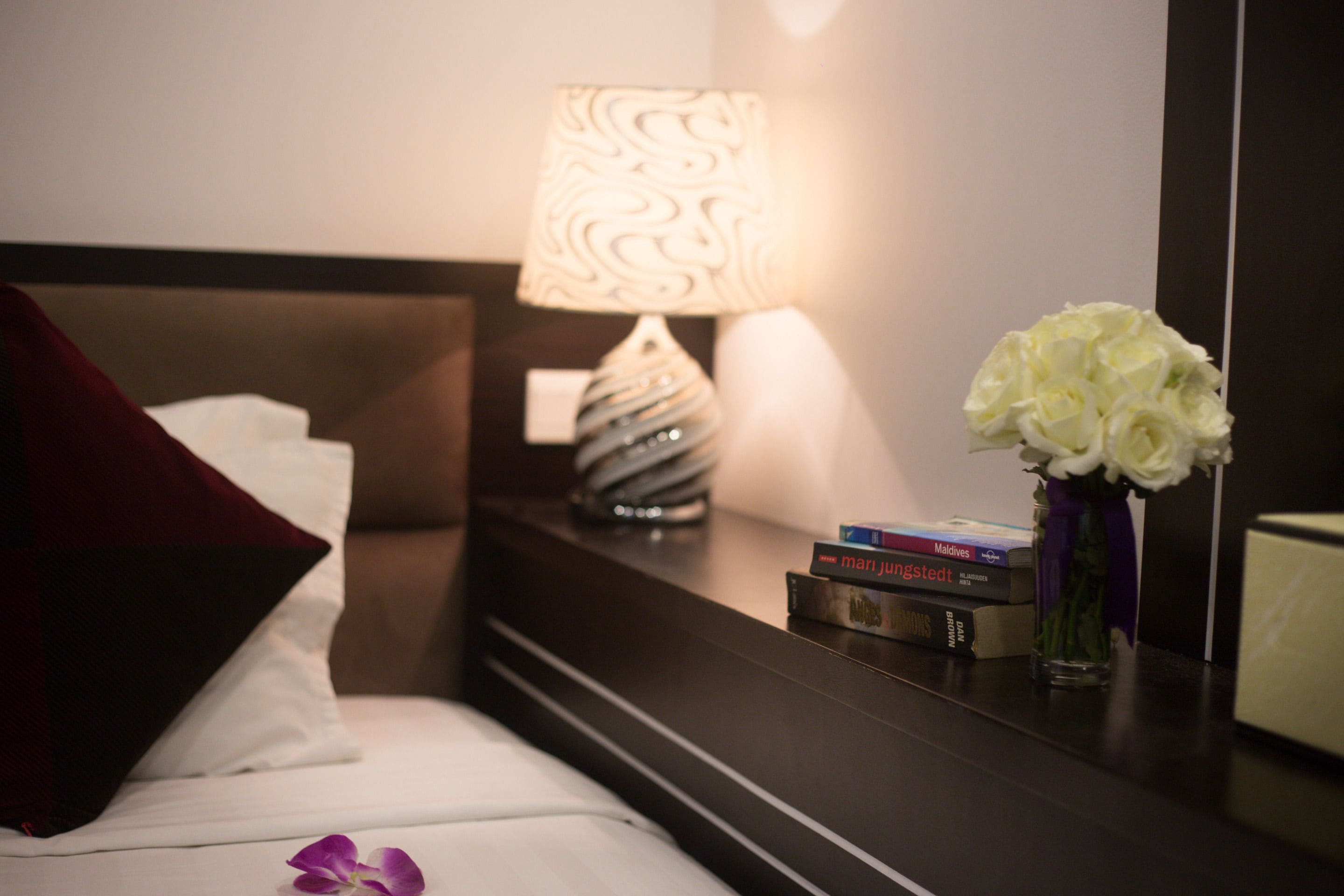 A neatly made bed with a white pillow and a red throw, a bedside table with a lamp, books, and a vase with white flowers.