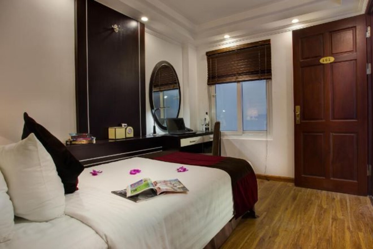 A bedroom with a bed, a desk, a chair, a large mirror, a window with blinds, and a wooden door.