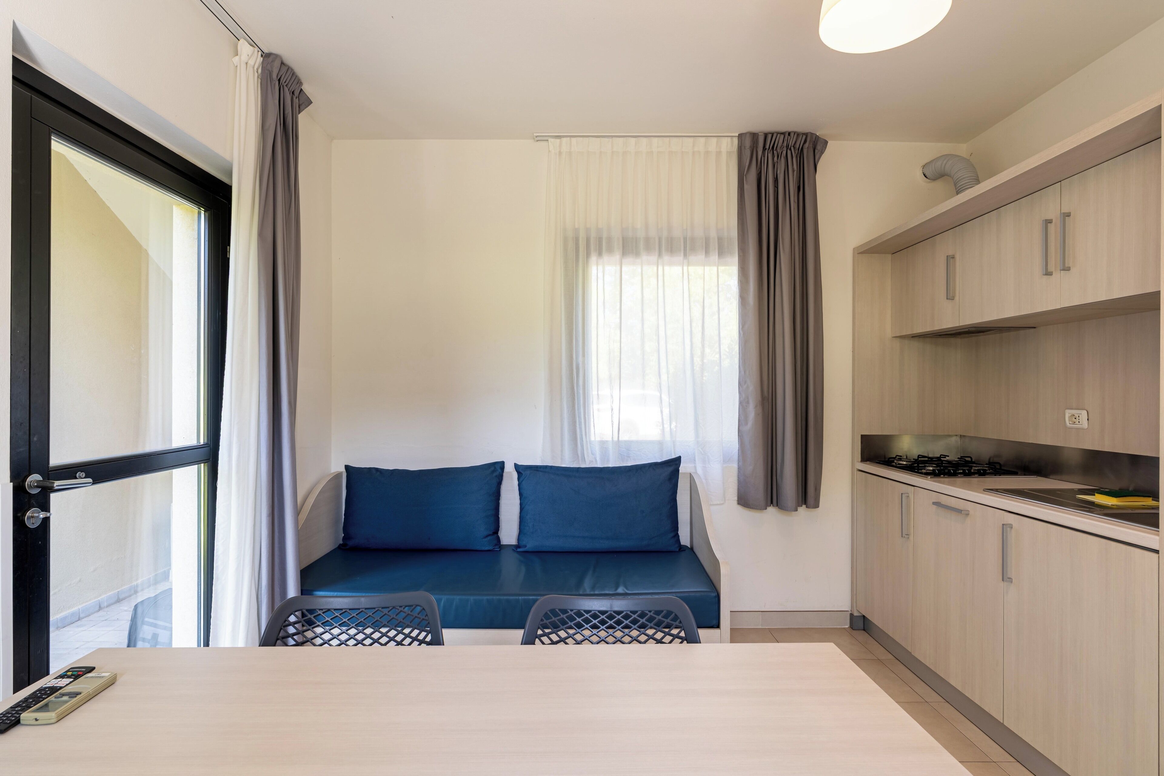 A compact room with a kitchenette, a dining area with a table and chairs, a blue sofa with cushions, and a window with curtains.